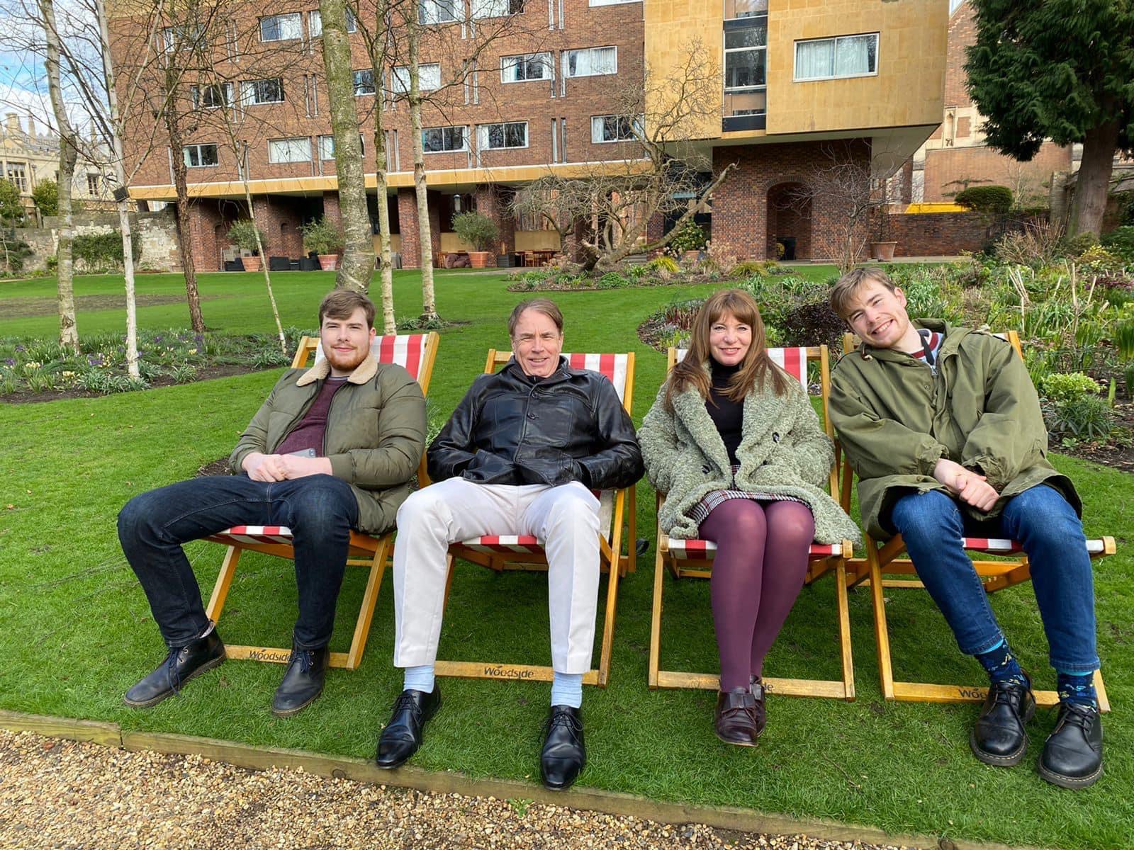 Four people sit smiling on deckchairs. They are sat on a green lawn, with trees and a brick building in the background. The people look relaxed and happy.