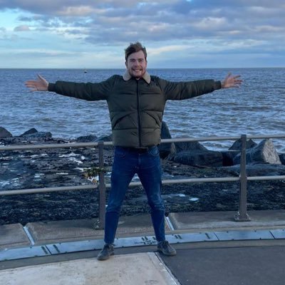 A man stands with his arms outstretched, making a star shape. He is smiling and wearing a green jacket and blue jeans. He is standing on a platform with the sea visible in the background/