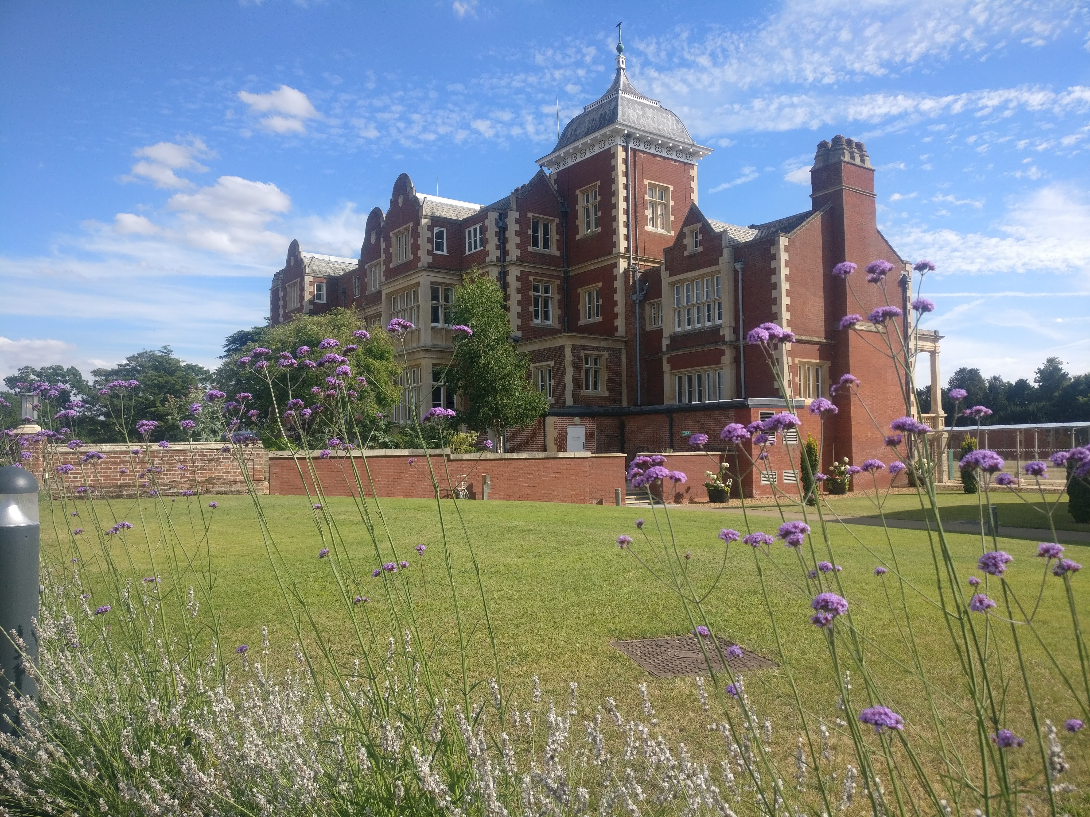 A grand redbrick building with purple flowers in the foreground and a well kept lawn