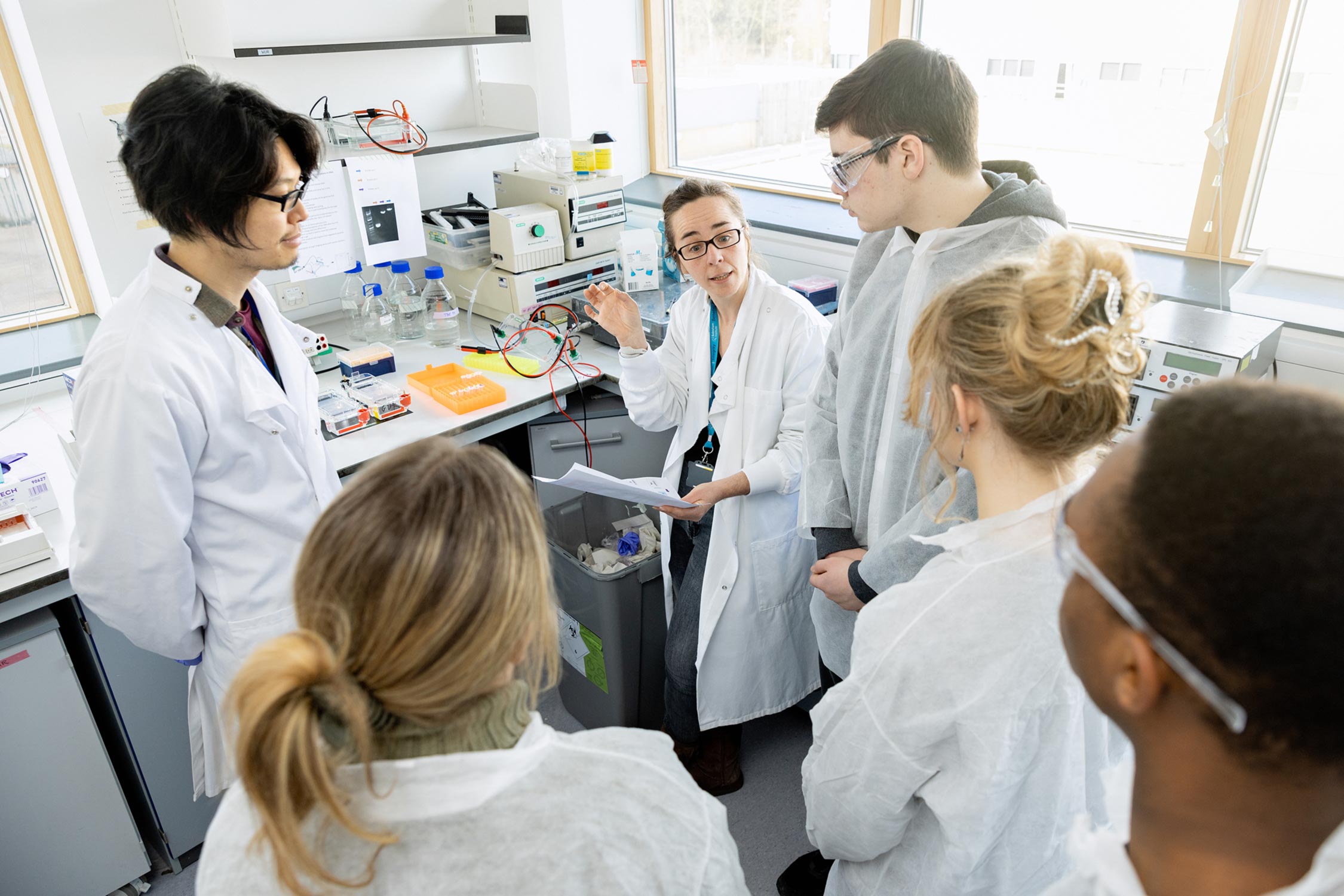 A Group Leader, sitting down and gesturing explains a task. A researcher and four students surround her, listening