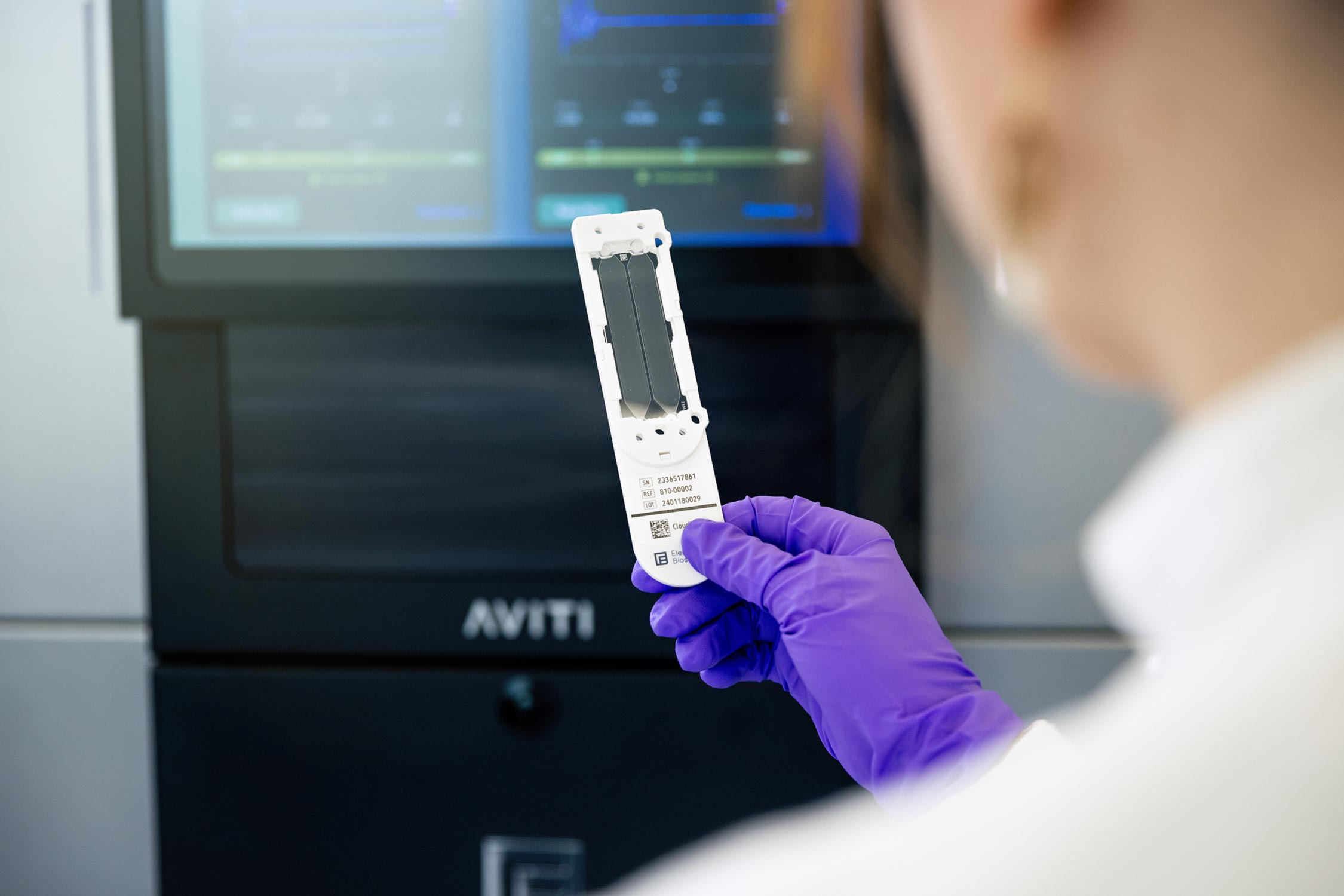 A technical specialist holds a flow cell for a next generation sequencer (AVITI sequencer shown in background).